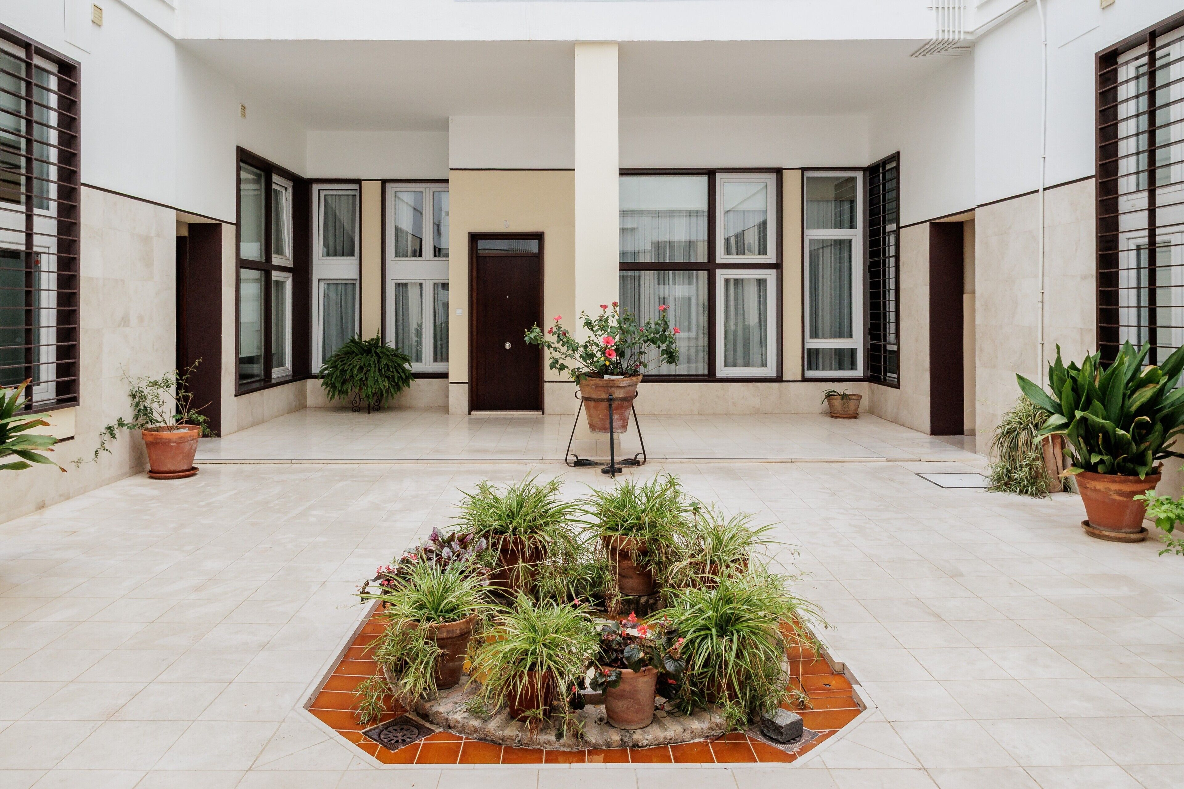 A spacious courtyard with potted plants arranged in a circular pattern, a central column, and large windows with blinds.