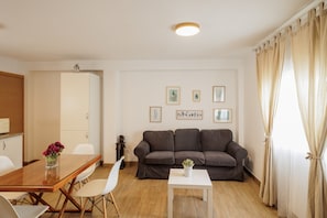 Una sala de estar moderna con un sofá, una mesa de comedor con sillas, una mesita blanca y una ventana con cortinas.