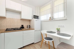 A modern kitchen with white cabinets, a gas stove, a microwave, and a refrigerator. There is a small table with two stools and a potted plant on the windowsill.