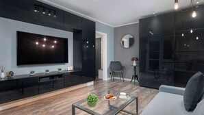 Un salón moderno con un televisor de pantalla plana grande, una mesa de centro de vidrio con objetos decorativos, un sofá gris y muebles de tono oscuro.