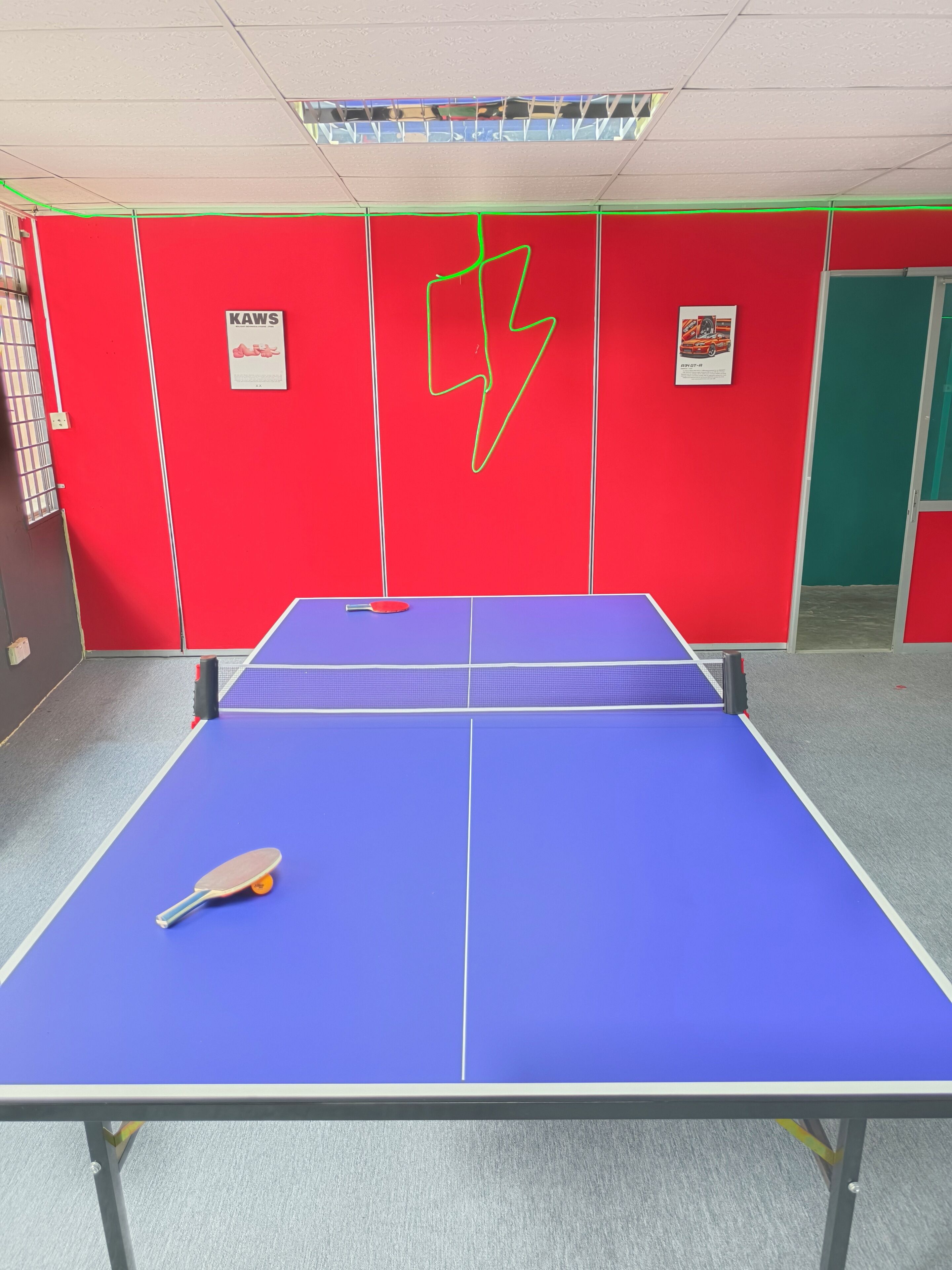 Una mesa de ping pong con una paleta, paredes rojas y dos carteles.