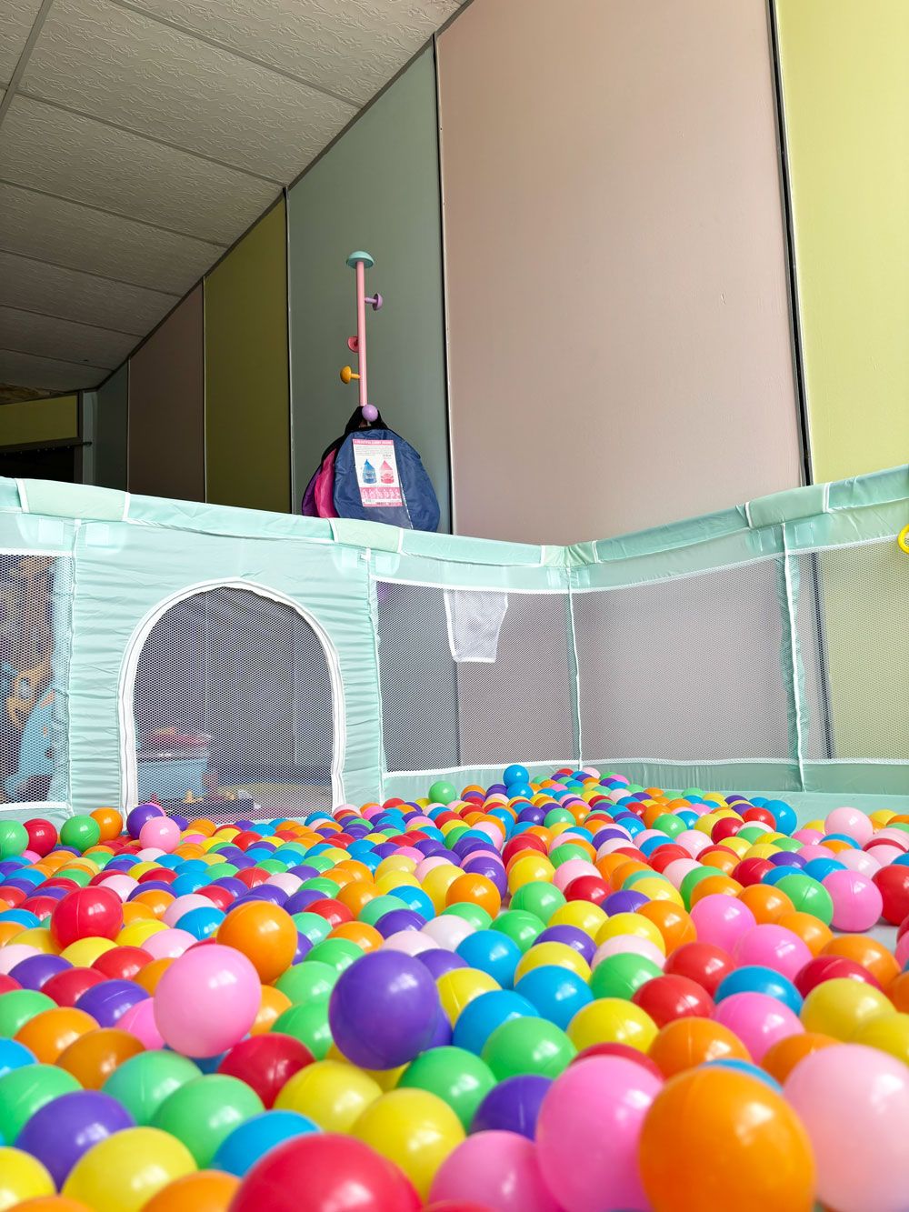 Een bällebak vol kleurrijke plastic ballen, een veiligheidshek en een roze en blauwe tas hangend aan een paal.