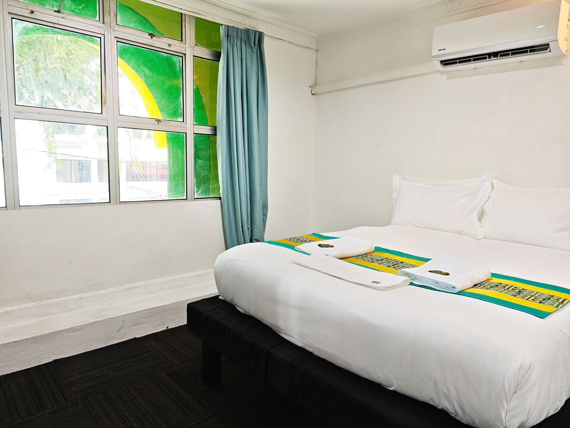 A hotel room with a bed, a bench, a window with curtains, and an air conditioning unit.