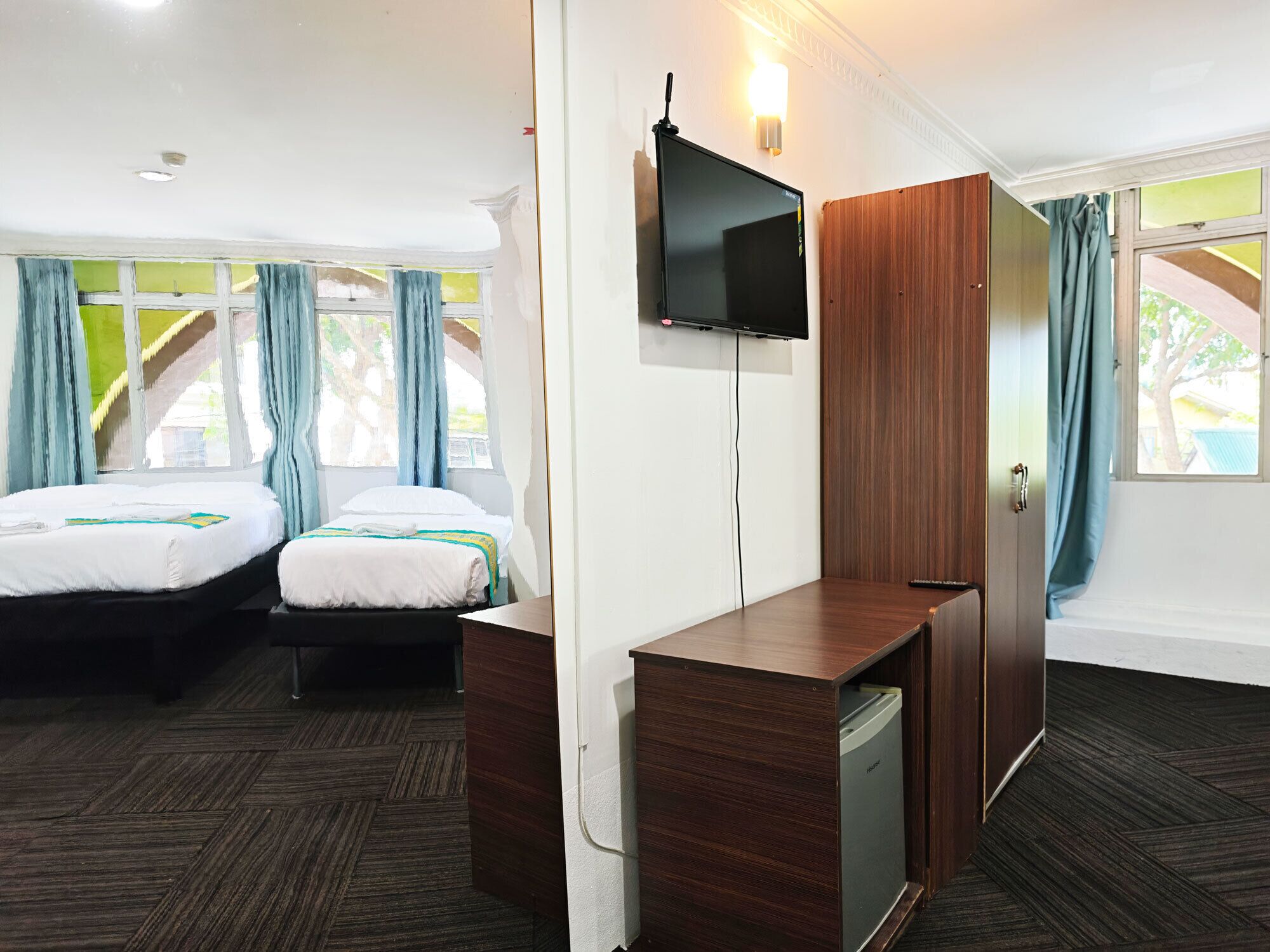 A hotel room with two beds, a TV mounted on the wall, a wooden wardrobe, and a small refrigerator.
