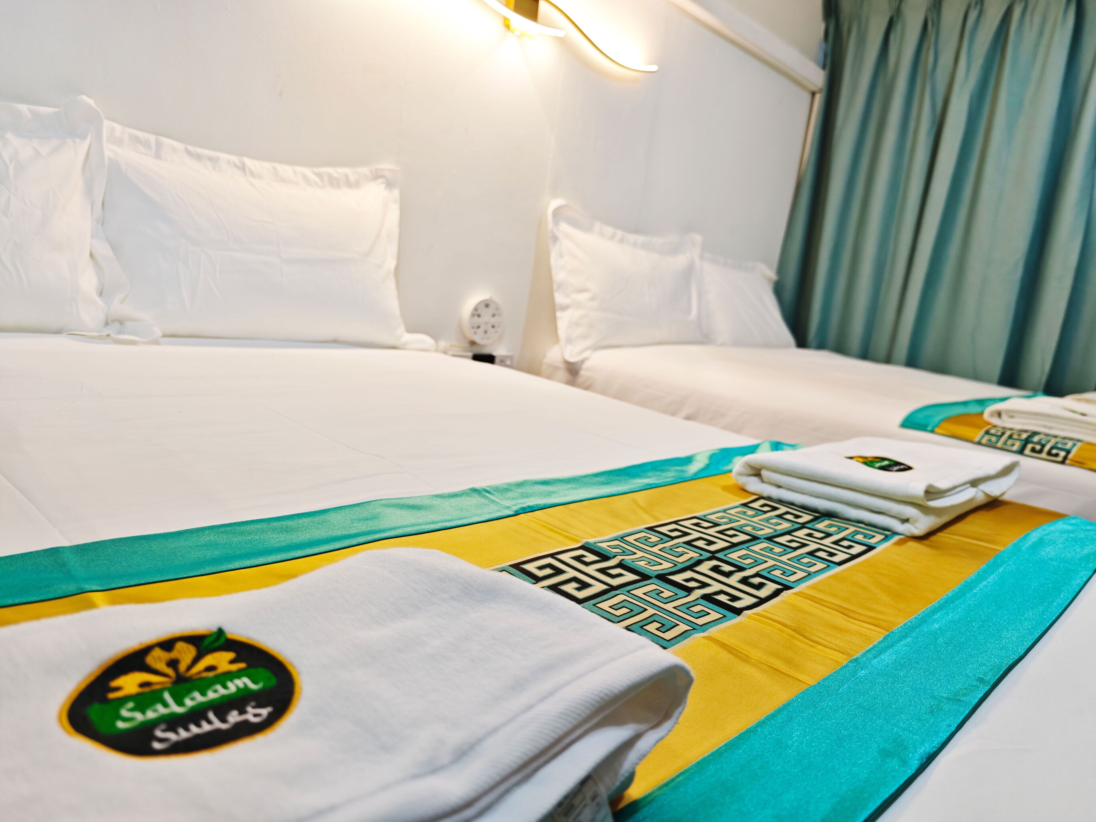 A hotel room with two beds, white bedding, a decorative bed runner, and a folded towel with a logo.