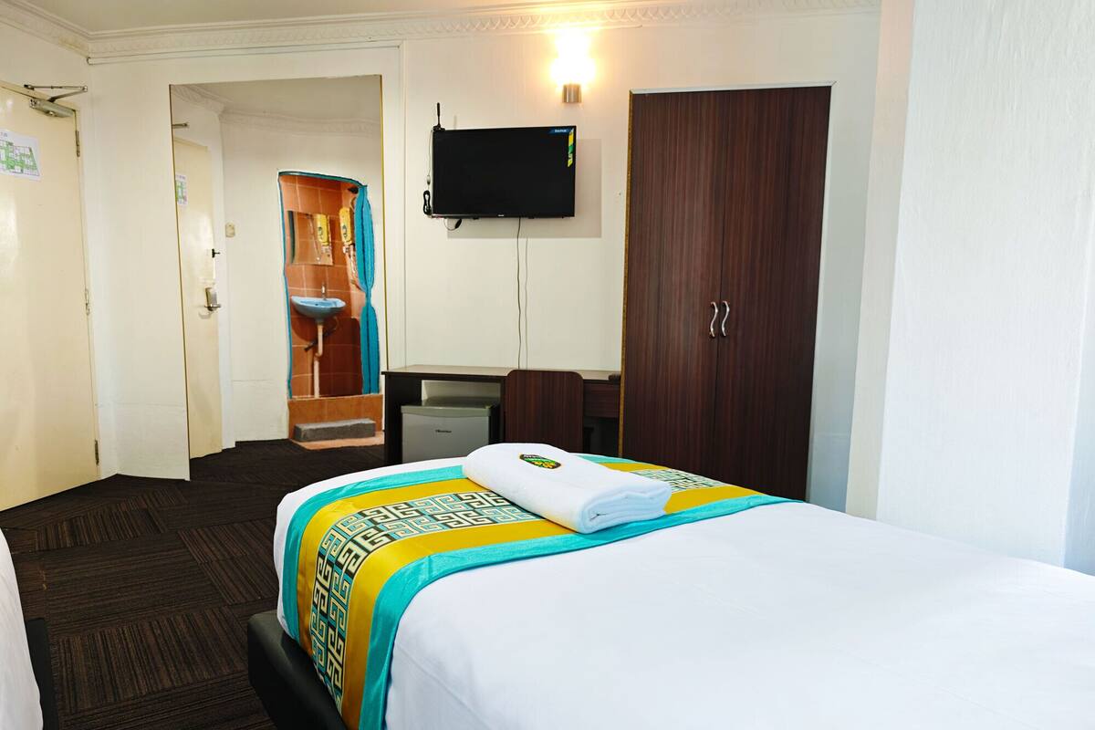 Quarto de hotel com cama, escrivaninha, televisão, guarda-roupa e banheiro.
