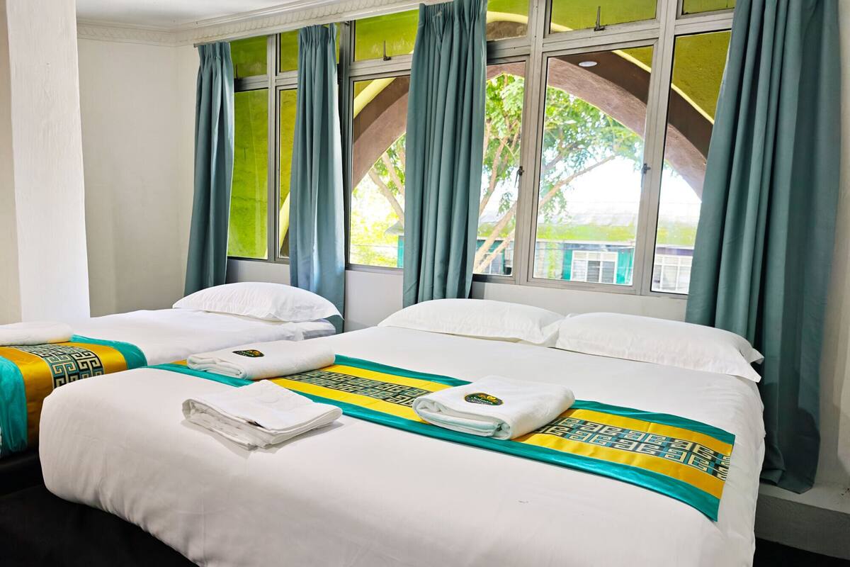 Quarto de hotel com duas camas, cada uma com roupa de cama branca e um cobertor listrado em verde e amarelo. Há dois jogos de toalhas em cada cama. O quarto tem janelas amplas com cortinas azuis, e a vista inclui vegetação lá fora.