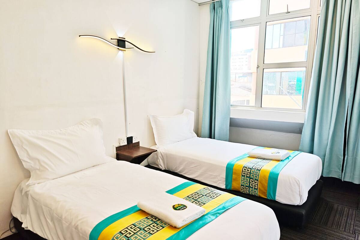 Quarto de hotel com duas camas, mesa de cabeceira, luminária na parede e janela com cortinas.