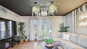 Una sala de estar moderna con un sofá modular, una mesa de centro y un televisor. Hay plantas en macetas, un candelabro y un tapiz de macramé.