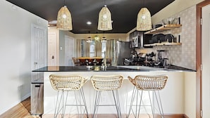Una cocina con isla central, tres taburetes de mimbre, lámparas colgantes y un comedor con mesa y sillas.