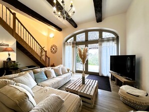 Una sala de estar con un sofá modular, una mesa de centro de madera, un televisor, una escalera y un amplio ventanal de arco.