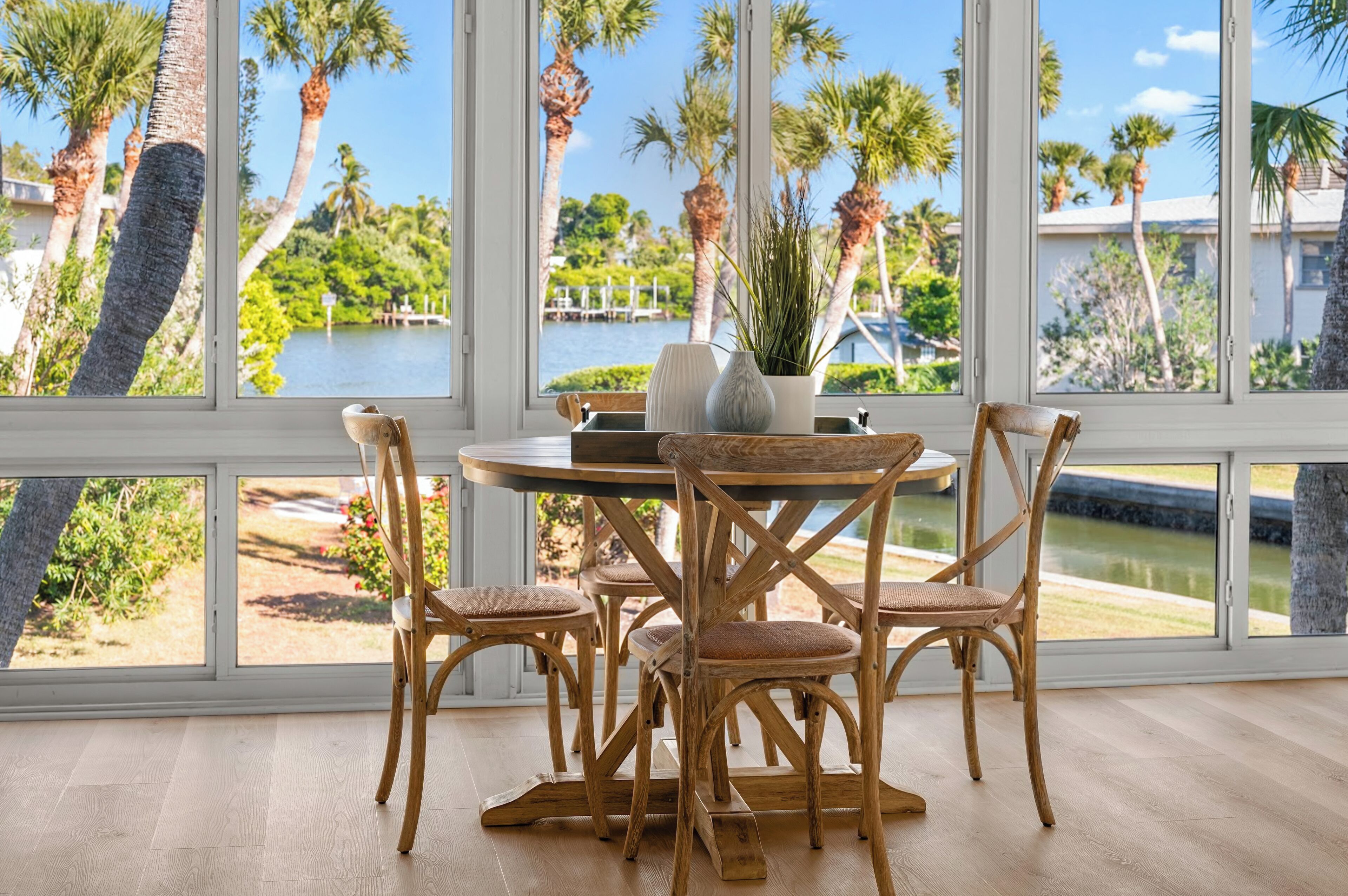 A dining area with a round table and four wicker chairs, large windows with a view of palm trees and a body of water.