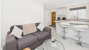 A modern living room with a grey sofa, two white bar stools, and a small round table with a vase.