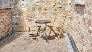 A small outdoor seating area with a round table and two wooden chairs, set against a stone wall.