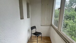 Une petite pièce étroite aux murs blancs, dotée d’un plancher en bois et d’une seule chaise noire. Une fenêtre se trouve sur un des murs, et une petite étagère avec un luminaire est fixée au mur.