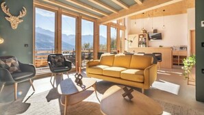 Amplia sala de estar con sofá, sillas y una mesa de comedor. Ventanas grandes ofrecen vistas a montañas.