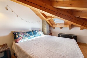Une chambre avec un plafond en pente, un lit recouvert d’une literie à motifs, une table de chevet en bois, une commode et une guirlande lumineuse.