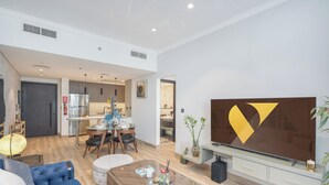 A modern living room with a large flat-screen TV, a dining area with a round table, and a kitchen in the background. - Vogue | Chaimaa Avenue JVC near Circle Mall (Dubai)