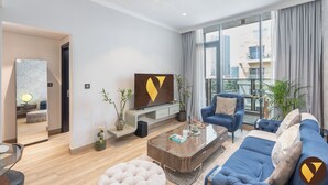A modern living room with a sofa, armchairs, a coffee table, and a TV. There is a large window with curtains, a plant, and a view of buildings outside. - Vogue | Chaimaa Avenue JVC near Circle Mall (Dubai)
