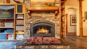 Une cheminée en pierre avec un feu, une bibliothèque remplie de livres et un tableau encadré au-dessus de la cheminée.