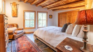 Une chambre avec un grand lit, un plafond en bois, deux fenêtres, une cheminée et un fauteuil en cuir.