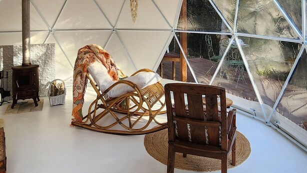 A tent with a geometric ceiling, a hanging chandelier, a rocking chair, and a view of the outdoors.
