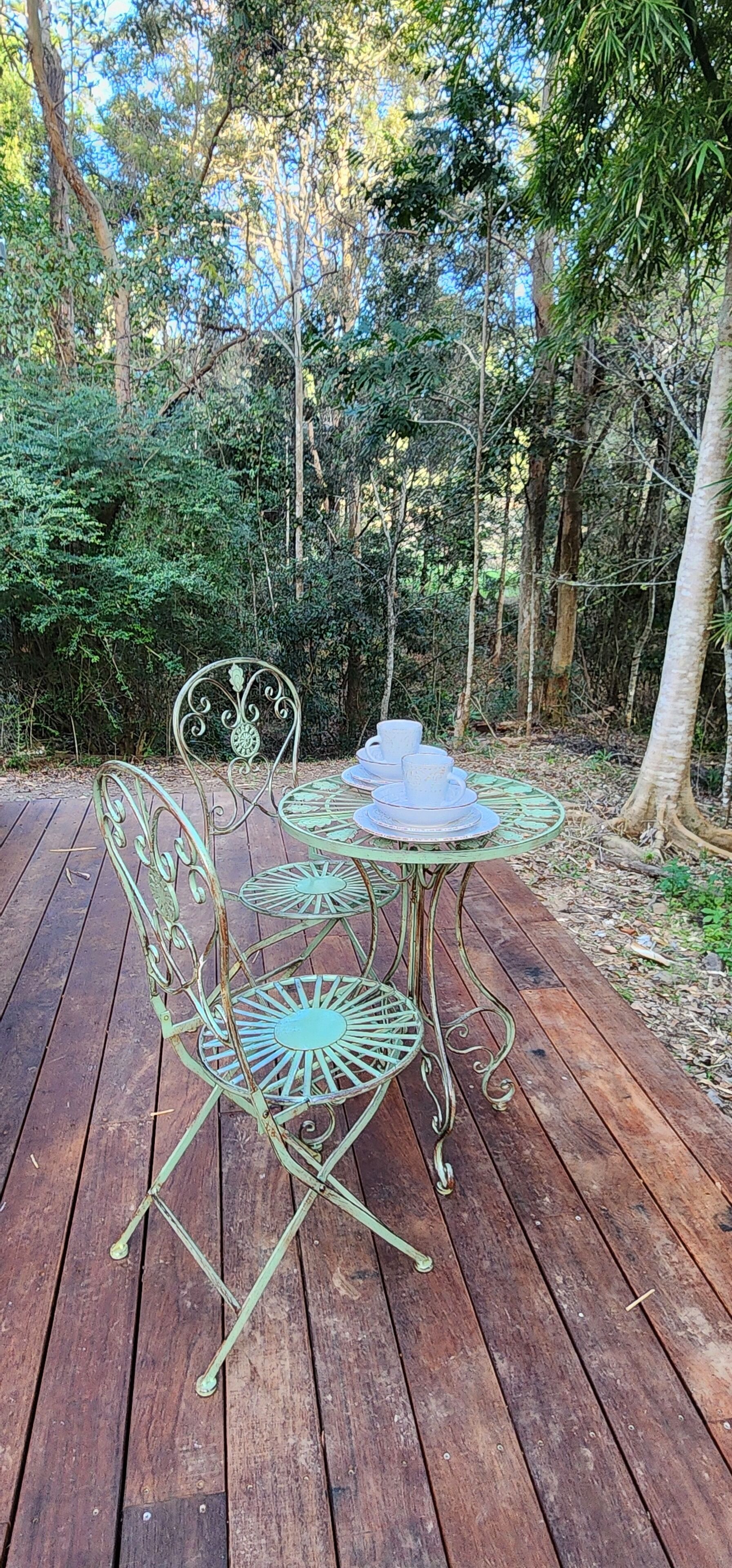 Een metalen buitentafel met stoelen op een houten terras, omgeven door dicht bos.