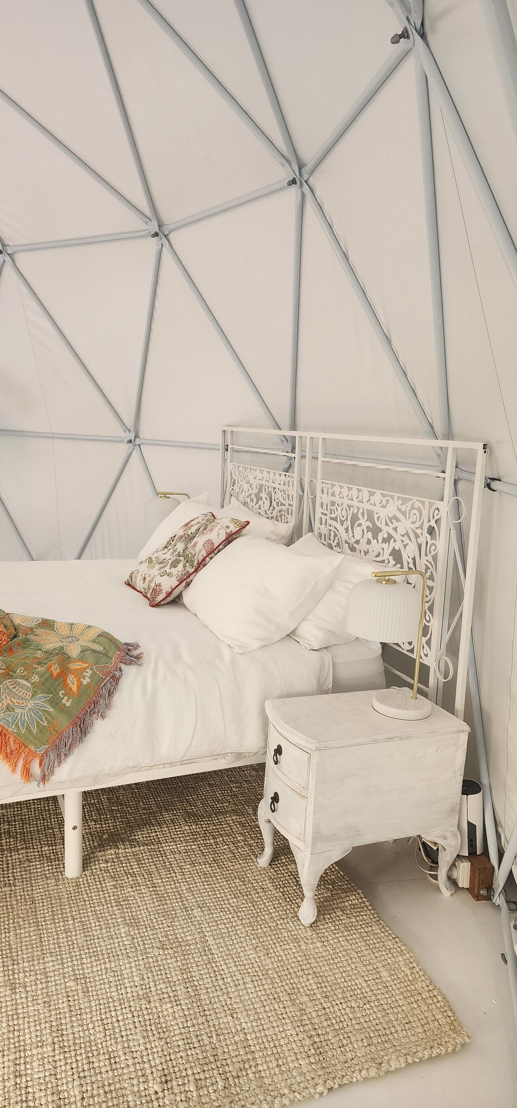 A bed with a white metal frame and a matching nightstand, a woven rug, and a geometric-patterned ceiling.