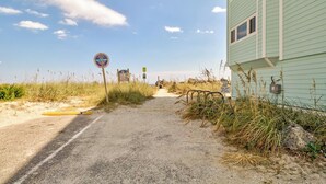 Una carretera de acceso a la playa con un letrero que indica 'Pago para Estacionar', un letrero que dice 'No Transgredir Dunas y Césped Marino', y un edificio de fachada verde claro.