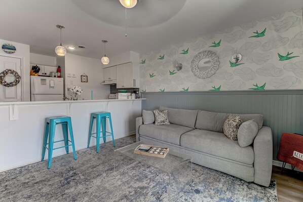 A modern kitchen with white cabinets, a grey sofa, and teal stools.