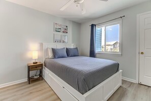 A bedroom with a bed, a nightstand, a ceiling fan, a window with curtains, and wall art.