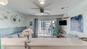 Una habitación moderna con tocador blanco, ventilador de techo, zona de comedor con sillas y vistas al balcón exterior.