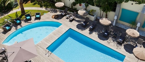 A large outdoor swimming pool with multiple lounge areas and umbrellas, surrounded by greenery and a white wall.