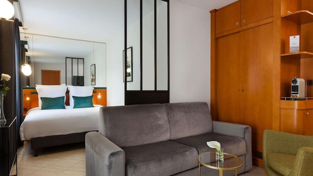 Une chambre d’hôtel moderne avec un canapé gris, une petite table ronde, un fauteuil vert, un lit avec une literie blanche et des meubles en bois.