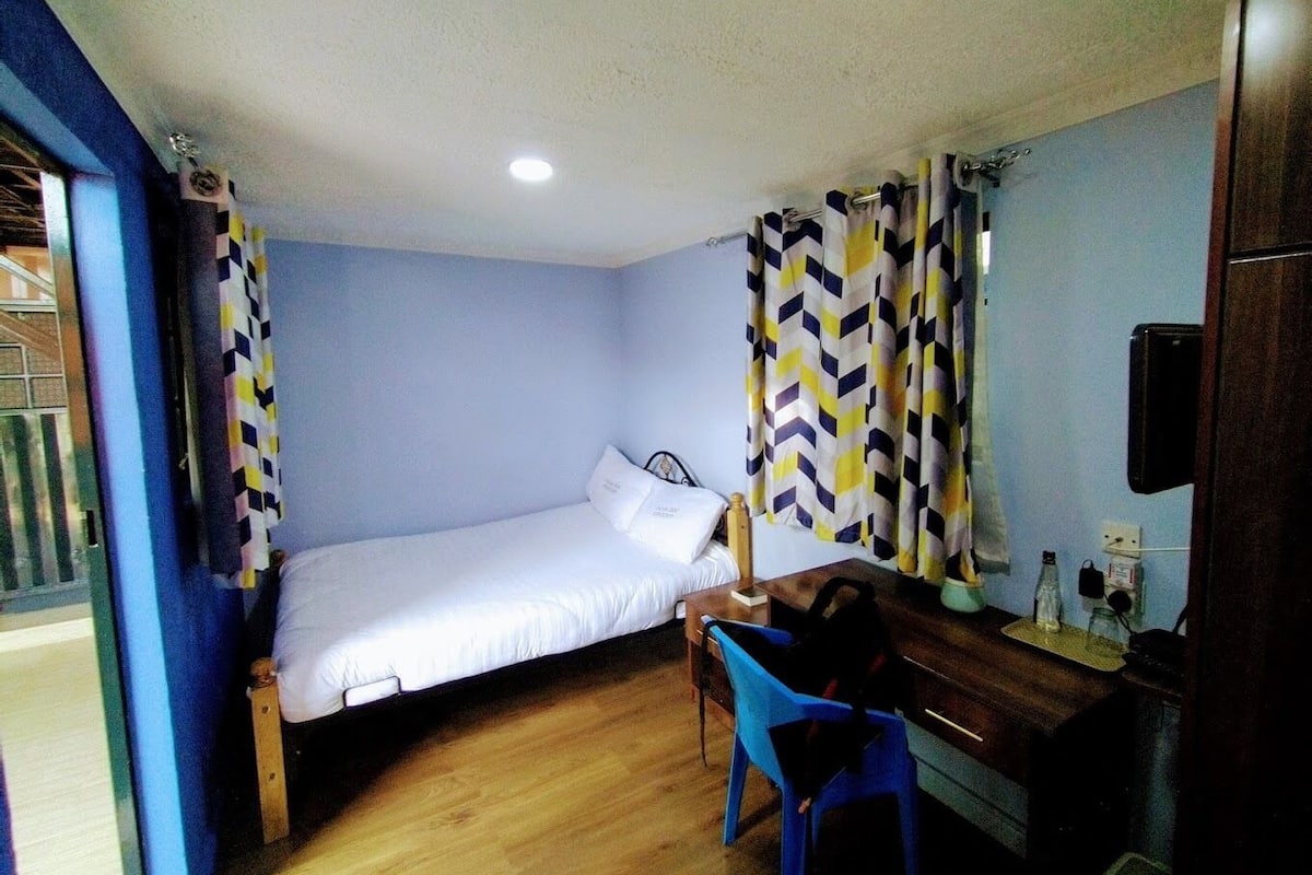 Ein kleines Zimmer mit einem Einzelbett, einem Schreibtisch, einem Stuhl, einem Spiegel und einem Fenster mit Vorhängen.