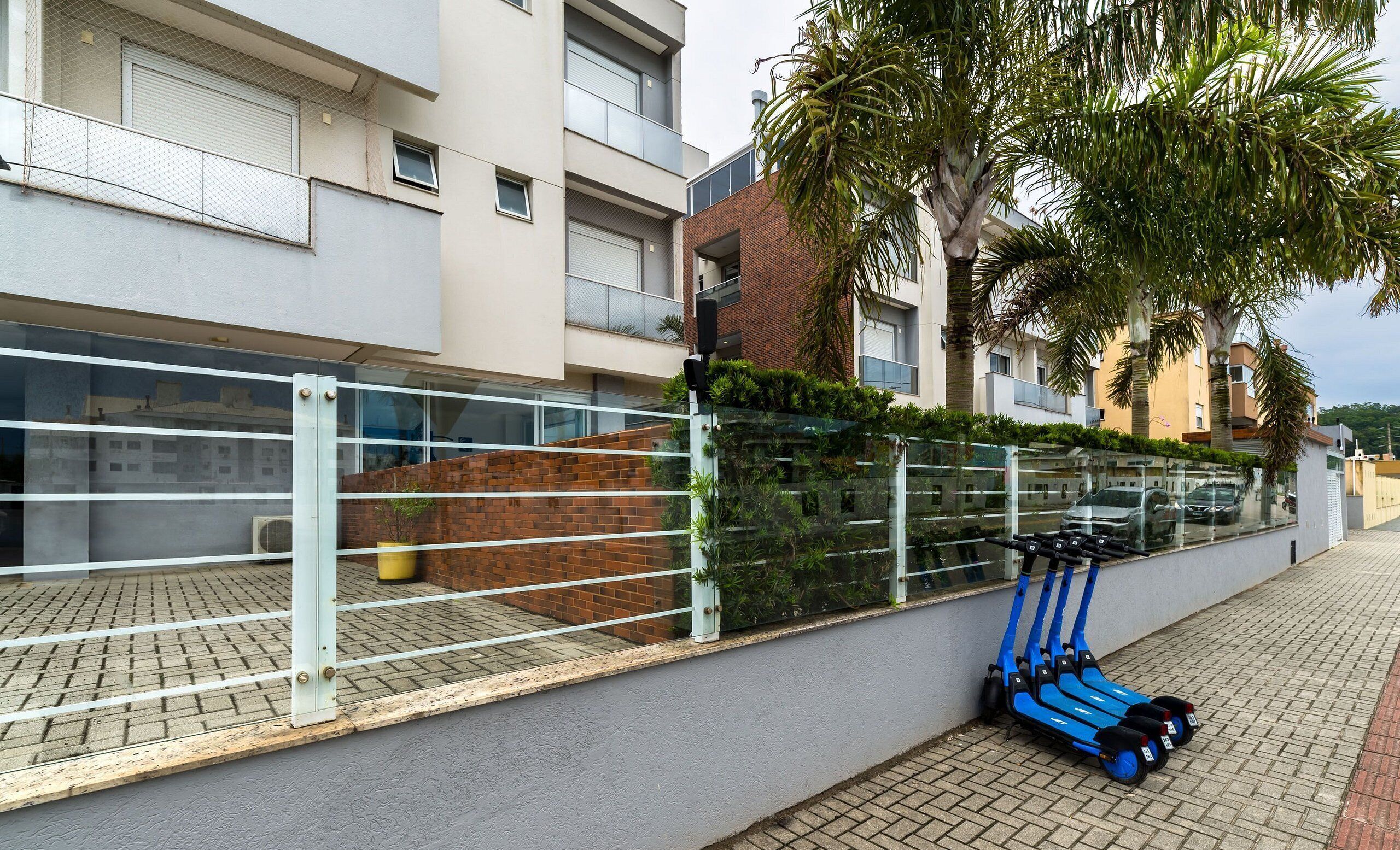 Un immeuble d’appartements moderne, avec une façade en briques, une clôture en verre et une rangée de trottinettes électriques bleues garées à l’extérieur.