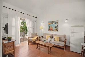 A living room with a sofa, a coffee table, a TV, and a refrigerator. There is a window with curtains, a lamp, and a wall with a picture.
