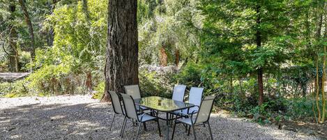 An outdoor dining area with a round table and four chairs, surrounded by trees and gravel.
