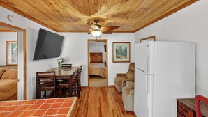 A compact room with a wooden ceiling, a flat-screen TV mounted on the wall, a dining table with chairs, a bed with a beige bedspread, a white refrigerator, and a small cabinet. - Rouge Cobb Mountain Hotel - Cabin 5 (Cobb)