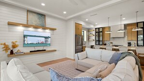 A modern living room with a large flat-screen TV, a sofa, and a dining area with chairs and a kitchen in the background. - Miramar House, 120 E. Constellation 6 Bedroom Home (South Padre Island)