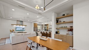 A modern kitchen with white cabinets, wooden shelves, and a dining area with a wooden table and white chairs. - Miramar House, 120 E. Constellation 6 Bedroom Home (South Padre Island)