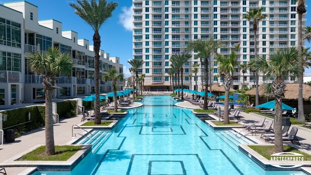 A hotel with a central swimming pool surrounded by lounge chairs and palm trees, with a multi-story building in the background. - Sapphire Condo & Spa Unit # 406 3 Bedroom Condo (South Padre Island)