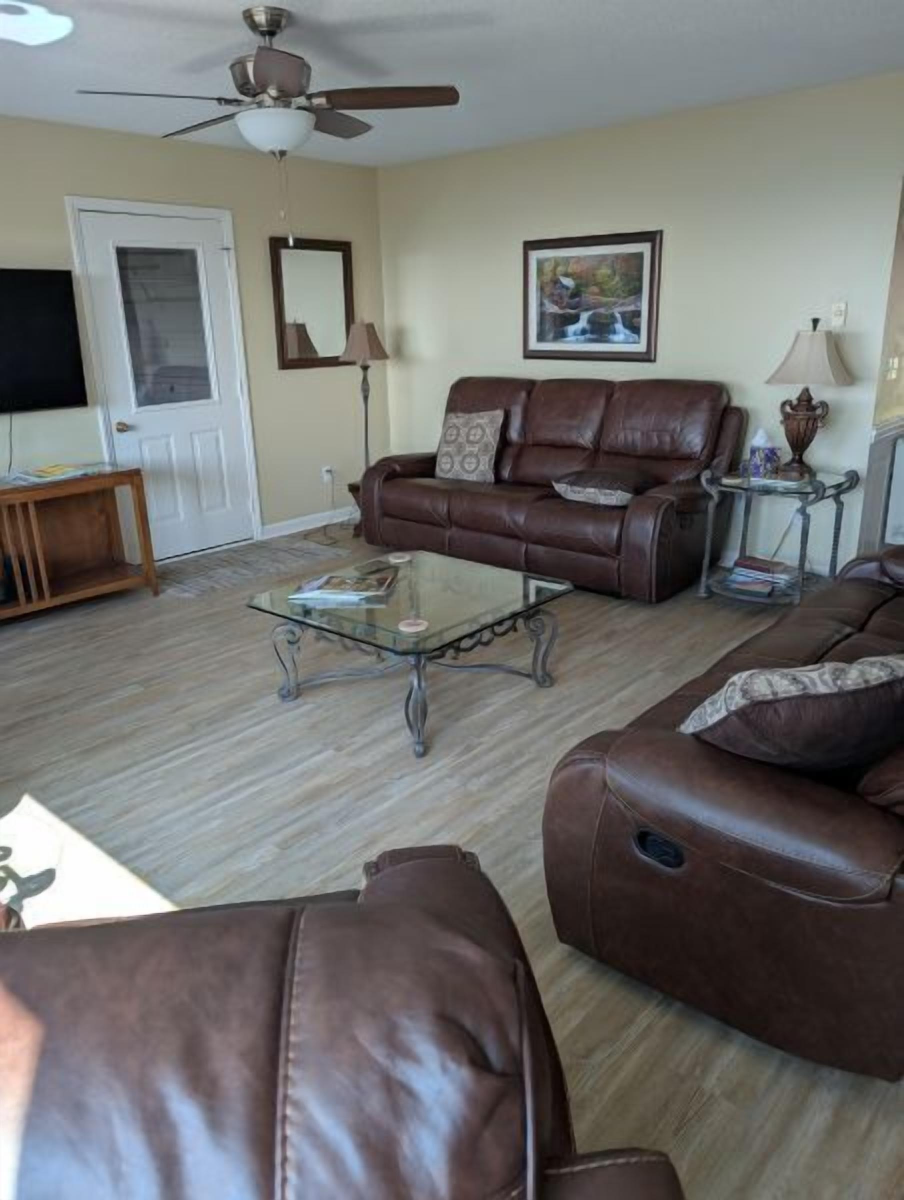 A living room with a brown leather sofa, a glass coffee table, a ceiling fan, a door, and a framed picture on the wall.