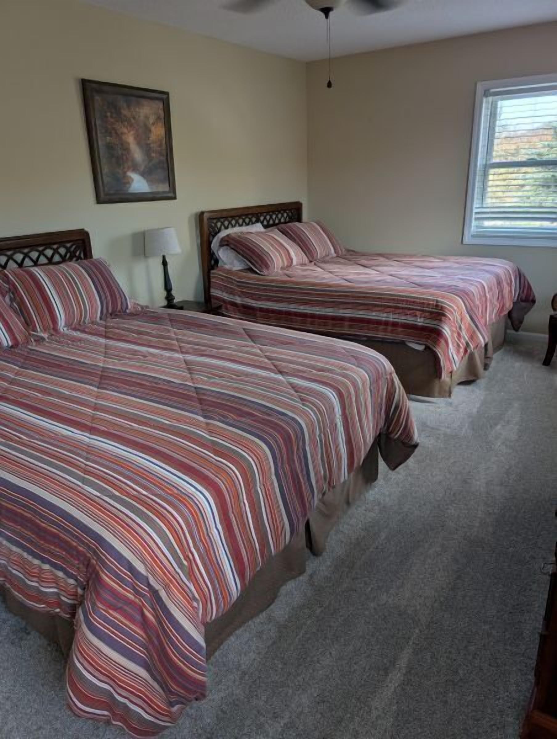 A bedroom with two beds, a ceiling fan, a window with blinds, and a framed picture on the wall.