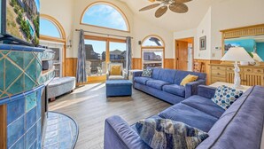 A spacious living room with a large sofa set, a dining area with a wooden table, and a balcony with a view of the surrounding buildings. - Oceanside, Dog-Friendly Home in Rodanthe with Private Pool and Hot Tub (Rodanthe)