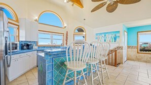 A kitchen with a blue tiled island, white bar stools, and a view of the sea through large windows. - Oceanside, Dog-Friendly Home in Rodanthe with Private Pool and Hot Tub (Rodanthe)
