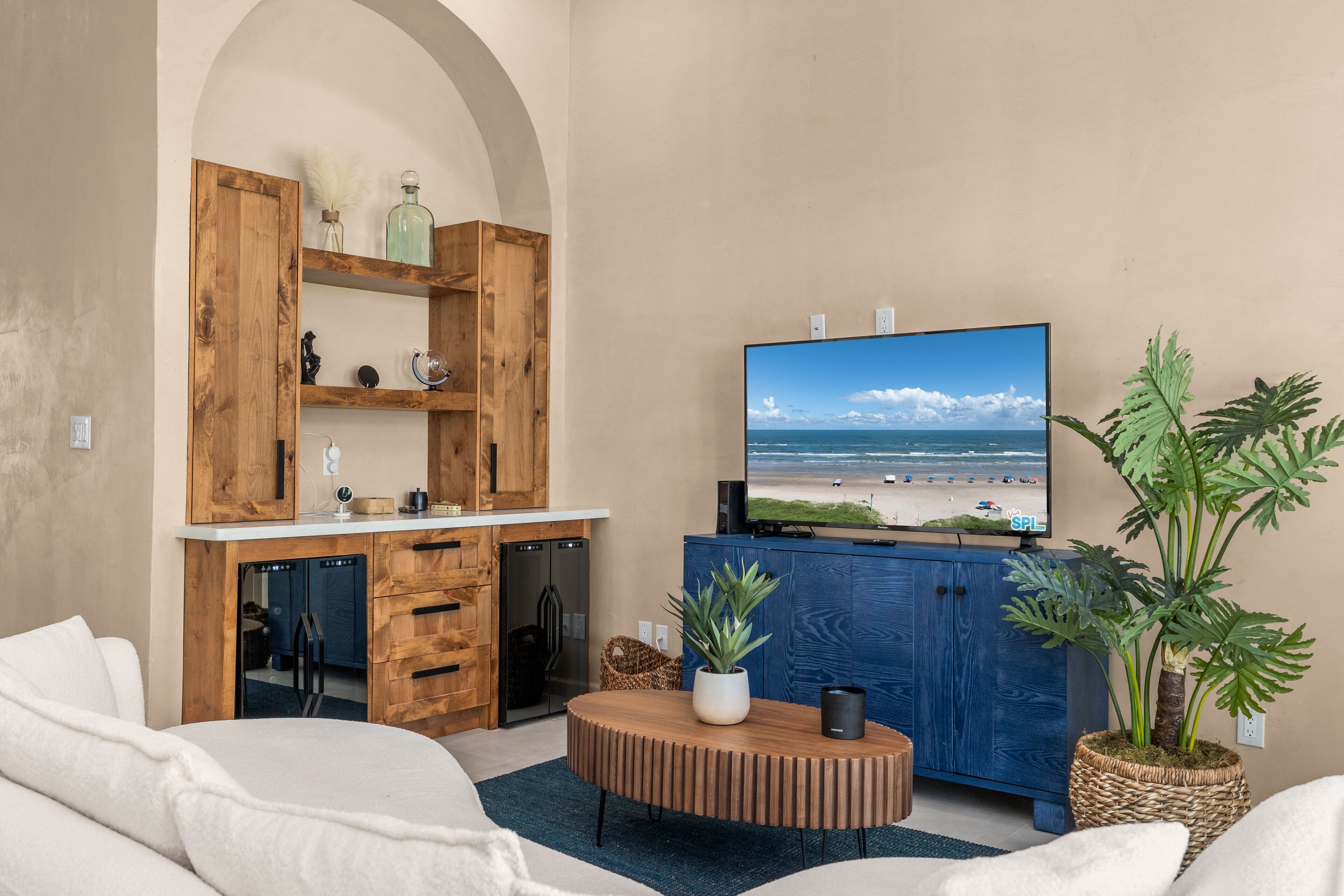 A modern living room with a large flat-screen TV, wooden furniture, and a view of the beach.