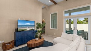 A modern living room with a large TV, a blue cabinet, a white sofa, and a view of the beach through the window.