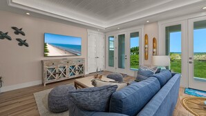 Ein geräumiges Wohnzimmer mit einer großen Couch, einem Fernseher und Blick auf den Strand durch Glastüren.