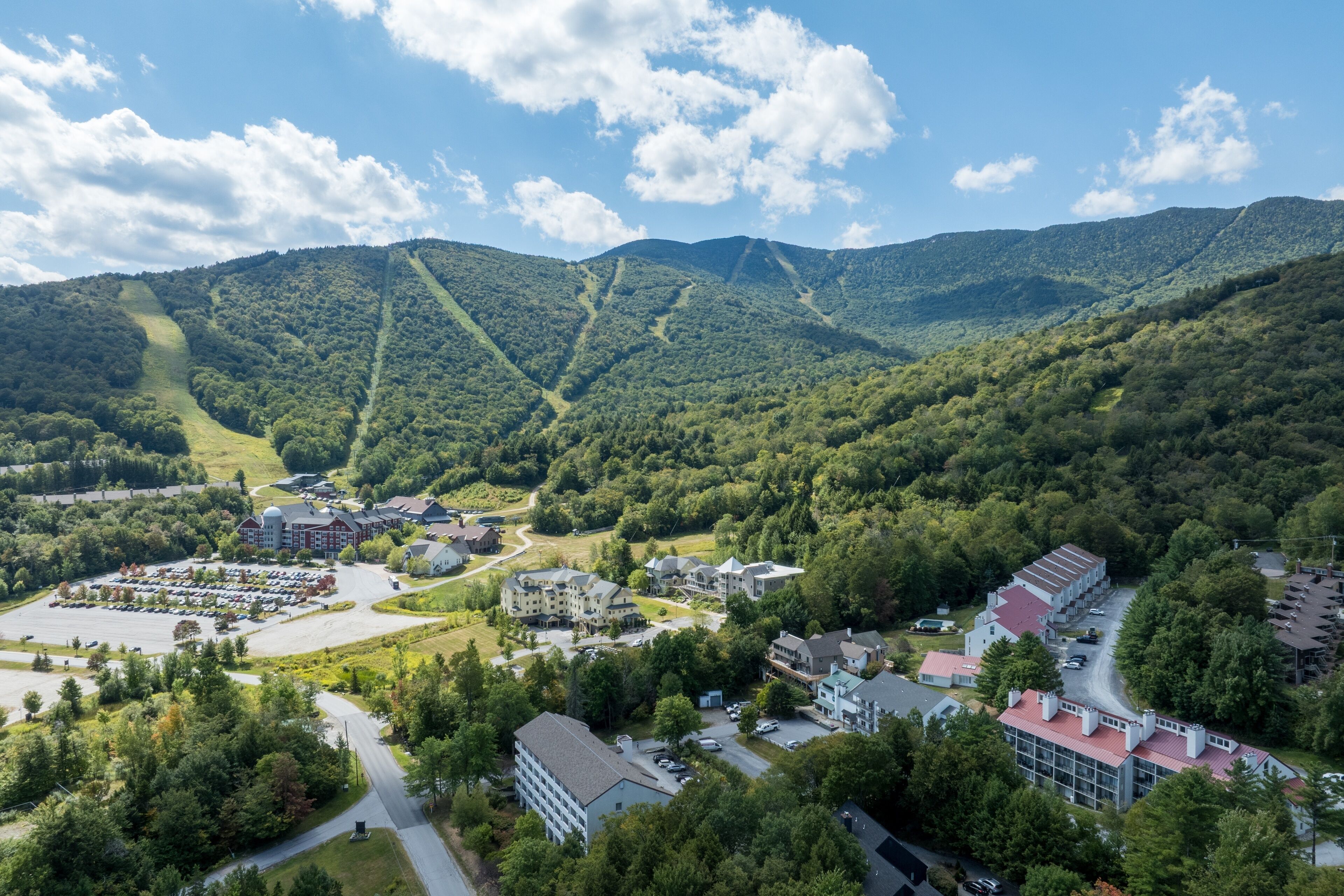 An aerial view of a resort with multiple buildings, a parking lot, and surrounding forested hills.